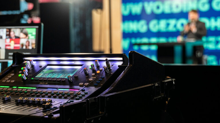 A broadcast mixing dashboard with a television host in the background